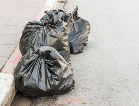 Two large black garbage bags filled with waste are stacked on an asphalt pavement near the edge of a concrete sidewalk in an outdoor urban setting. The bags are made of thick, shiny plastic with visible folds and creases, and appear to contain mixed rubbish. They are positioned close to a red and white painted curb, which separates the roadway from the pavement. A small metal litter picker or grab tool is leaning against one of the bags, suggesting they have been recently filled or are awaiting collection. The environment suggests a typical street scene in Vauxhall, within the postcode area associated with south London, and is indicative of waste removal or rubbish clearance activities often handled by local rubbish removal services such as Office Clearance Vauxhall. The background shows a plain, grey asphalt surface with no other objects or visible landmarks, emphasizing the focus on the waste bags ready for disposal or collection.