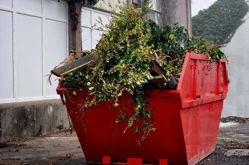 A large red skip bin filled with green foliage, small branches, and some wooden pieces, positioned on a paved surface outdoors next to a white wall with window panes. The skip has a slightly open lid on the right side, revealing the mixed contents inside. Surrounding the area is an urban environment, with concrete and brick elements visible underneath the foliage. The background includes a section of a building and a glimpse of some trees, suggesting the location is near a property in Vauxhall, London. The scene is lit with natural daylight, highlighting the texture of the debris and the vivid red color of the skip, typical of rubbish removal services such as those provided by Office Clearance Vauxhall, especially for garden or site clearance projects in the local area.