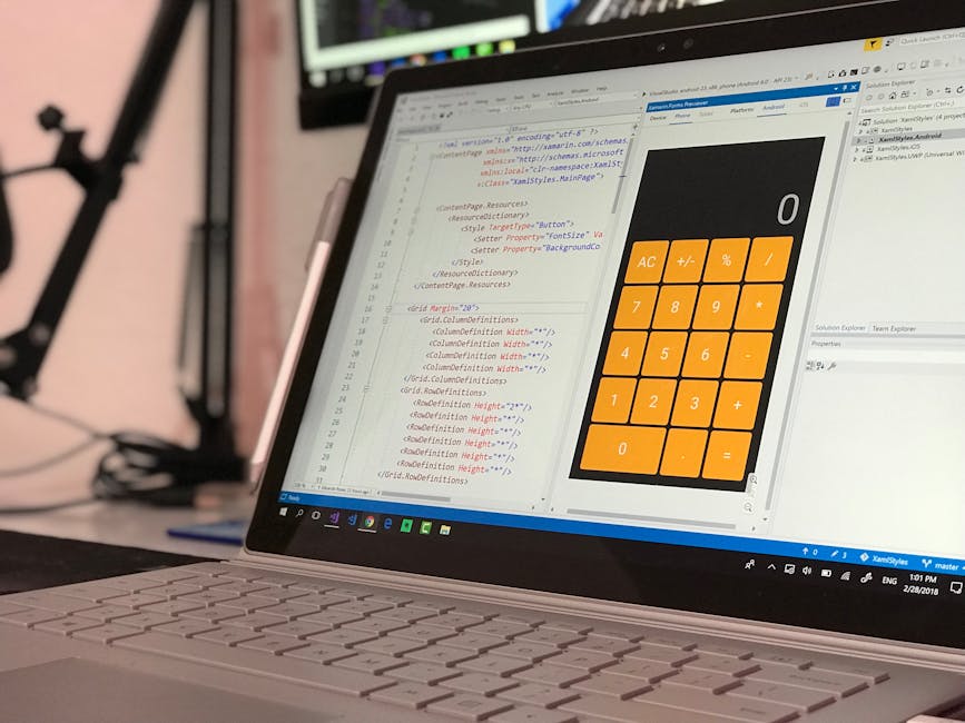 A close-up view of a laptop computer on a desk displaying a software development environment with lines of code in a text editor and a large on-screen calculator interface with orange buttons for basi