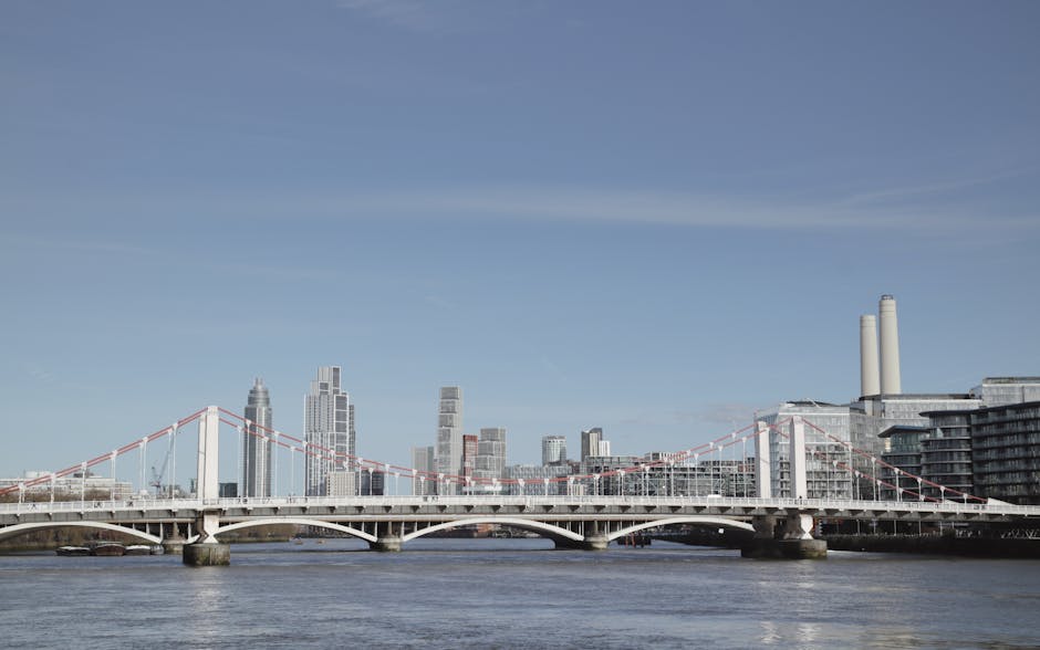 A wide view of the London skyline with a prominent suspension bridge spanning the River Thames in the foreground, supporting cables and a white arch structure. Behind the bridge, modern high-rise buil