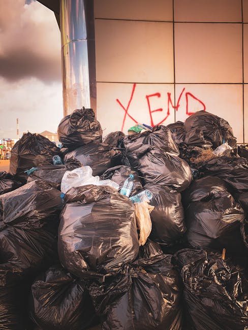 A large pile of black rubbish bags filled with waste materials is stacked against a modern building with a tiled exterior. Some bags appear to be slightly torn or bulging, revealing partially visible 