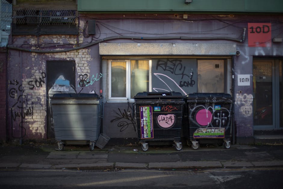A black wheeled rubbish bin labeled 'St. John's' positioned on a sidewalk beside a street at night, filled with various waste items including paper, cardboard, and plastic, with some items protruding